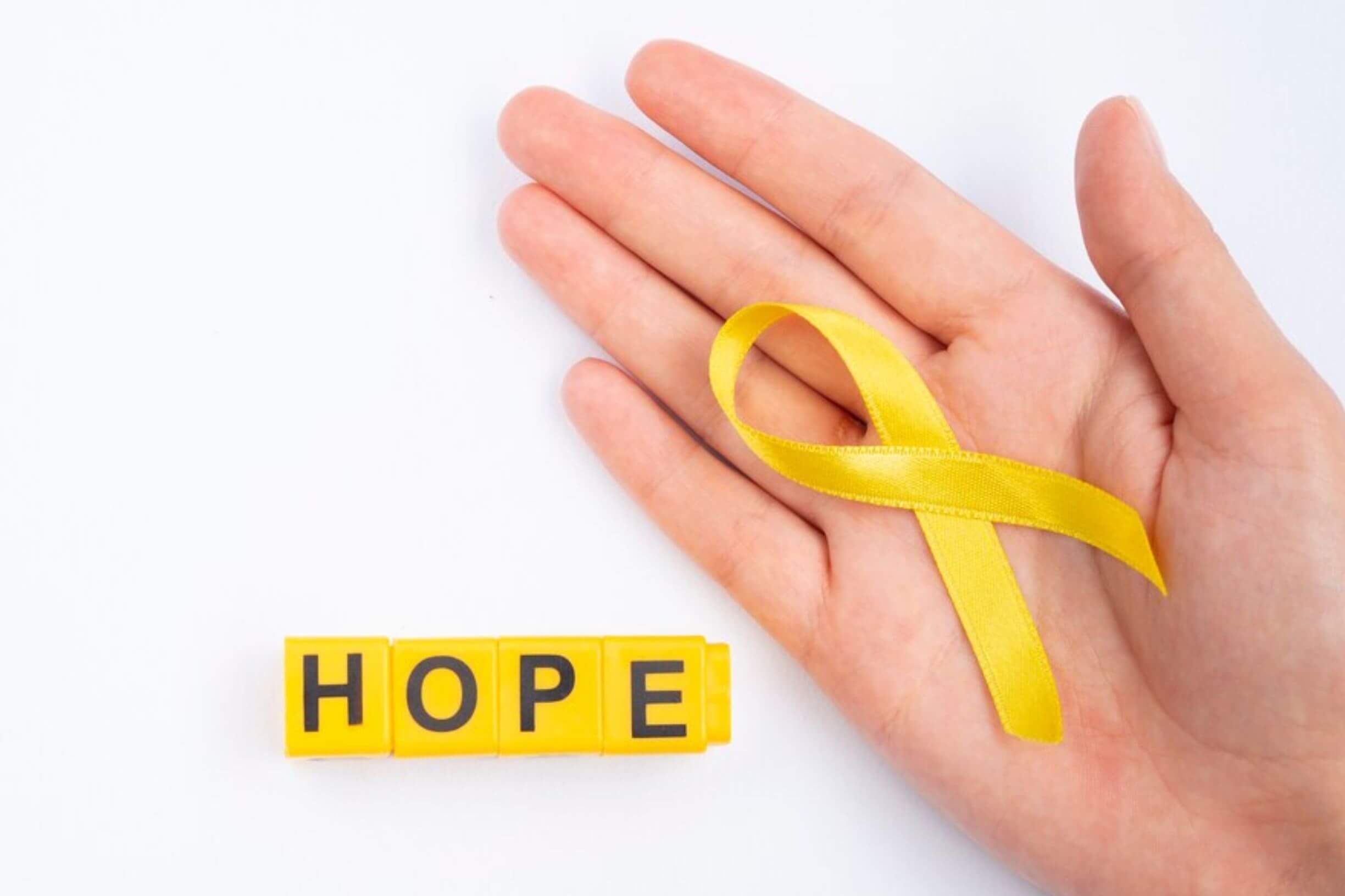 A person's hand holding a yellow ribbon, with the word "HOPE" spelled out in yellow blocks below.