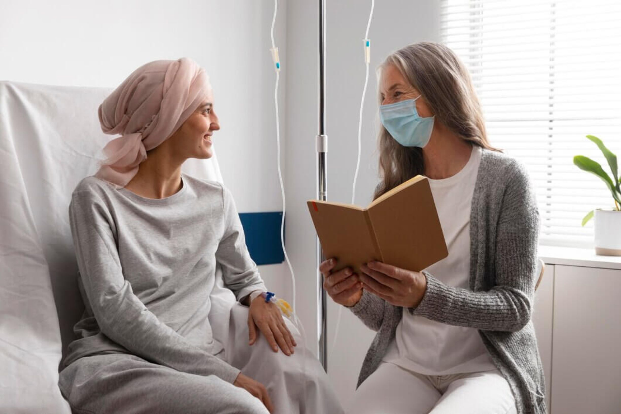 A cancer patient with a pink headscarf smiling while listening to a caregiver reading a book, both seated in a healthcare setting.